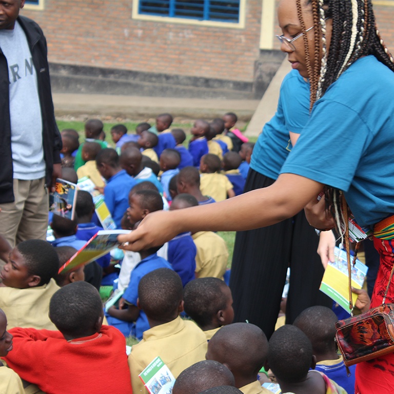 A woman hands out books to school children seated on grass